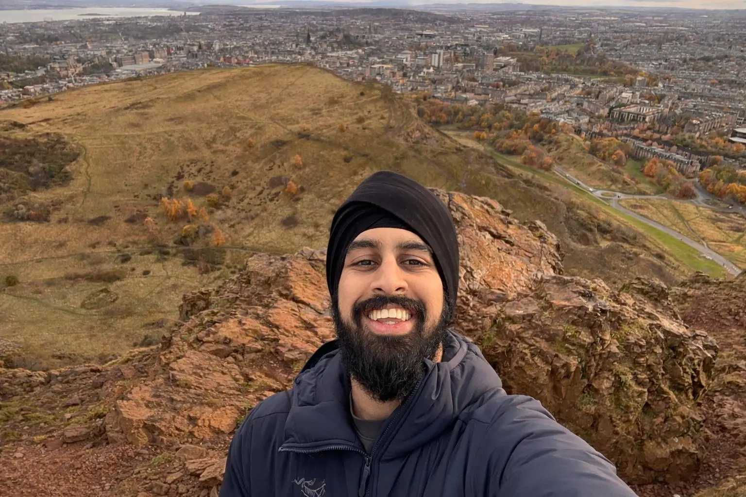 Nirmit smiling in a selfie on a rocky summit above Edinburgh